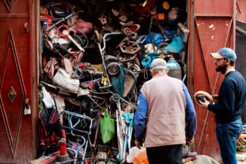 Two men inspecting a workshop with a cluttered collection of discarded items and tools.
