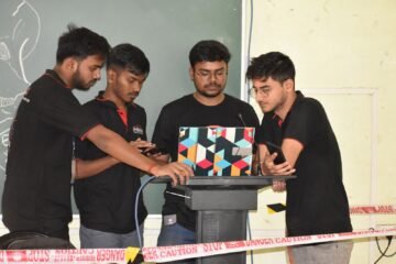 Four students work together on a laptop, preparing a presentation indoors by a chalkboard.