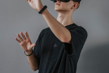 Young man in black shirt enjoying a virtual reality experience using a VR headset against a gray wall.