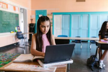 A focused young girl using a laptop for schoolwork in a bright classroom environment.
