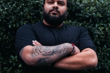 a man with tattoos standing in front of a brick wall